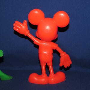 May include: A red plastic toy figurine of Mickey Mouse with large ears, wearing a red shirt and shorts. He is waving with his right arm.