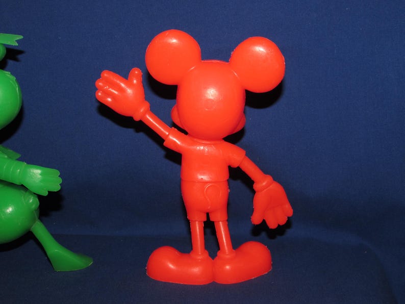 May include: A red plastic toy figurine of Mickey Mouse with large ears, wearing a red shirt and shorts. He is waving with his right arm.