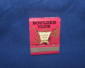 May include: A vintage matchbox with a pink background and the words "BOULDER CLUB" in bold black letters. The matchbox also features the text "SINCE 1929" and the locations "LAS VEGAS" and "NEVADA".