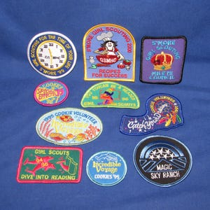 May include: A collection of embroidered patches with various designs and text. The patches feature themes like Girl Scouts, cooking, and travel. Colors include blue, green, red, and yellow. Some patches have text such as "S'more Girl Scouting" and "Dive into Reading."