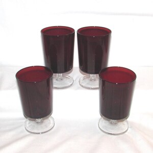 May include: A set of four matching drinking glasses featuring deep burgundy red bowls and clear glass stems with circular bases. The glasses have a cylindrical shape with a smooth finish, arranged in a two-by-two formation on a white surface.