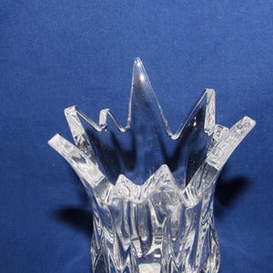 May include: Clear glass vase with a textured, ridged body and a starburst-shaped rim. The vase is transparent, allowing light to pass through, and sits against a blue background. The design is modern and decorative.
