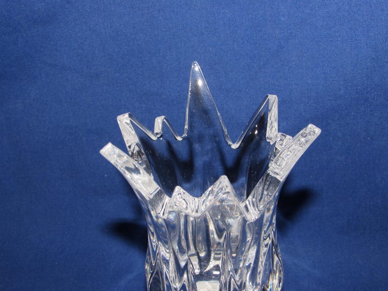 May include: Clear glass vase with a textured, ridged body and a starburst-shaped rim. The vase is transparent, allowing light to pass through, and sits against a blue background. The design is modern and decorative.