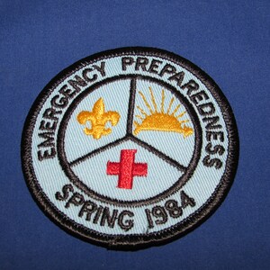 May include: A round, blue and black embroidered patch with the text "Emergency Preparedness Spring 1984". The patch features a red cross, a yellow fleur-de-lis, and a yellow sun.