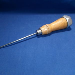 May include: A scratch awl featuring a long, slender, pointed metal shaft attached to a smooth, light-colored wooden handle. The handle is capped with a hexagonal metal nut at the end and features a metal ferrule where the shaft meets the wood.