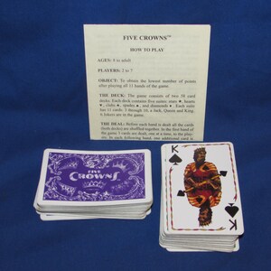 May include: A deck of Five Crowns playing cards with a purple and white card back design. The image includes a stack of cards, a card showing the King of Spades, and a sheet with game instructions. The game is for ages 8 to adult.