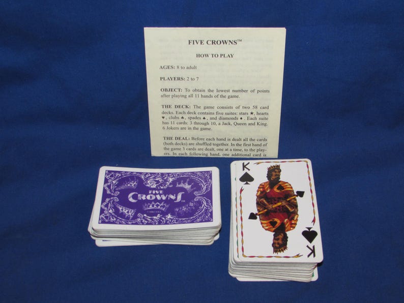 May include: A deck of Five Crowns playing cards with a purple and white card back design. The image includes a stack of cards, a card showing the King of Spades, and a sheet with game instructions. The game is for ages 8 to adult.