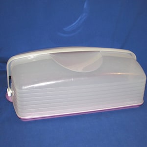 May include: A clear plastic cake carrier with a white handle and a purple base. The lid has a curved design and a clear top. The carrier is designed to hold a cake and keep it fresh.