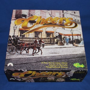 May include: A box for the Cheers board game, featuring a vintage illustration of the bar's exterior with a horse-drawn carriage in front. The box is yellow and gold with the word "Cheers" in a large, stylized font. The text on the box reads "A Fun Trivia Game About America's Favorite Neighborhood Tavern".