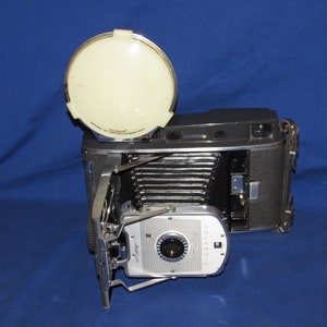 May include: A vintage Polaroid Land Camera with a flash attachment. The camera is silver and black with a black leather strap. The flash is white and round.