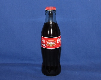 May include: A clear glass Coca-Cola bottle with a red and white label and a red cap. The bottle is filled with a dark liquid and has the Coca-Cola logo prominently displayed. The bottle is approximately 8 inches tall.