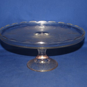 May include: A clear glass cake stand with a scalloped edge and a pedestal base. The stand has a delicate etched pattern.