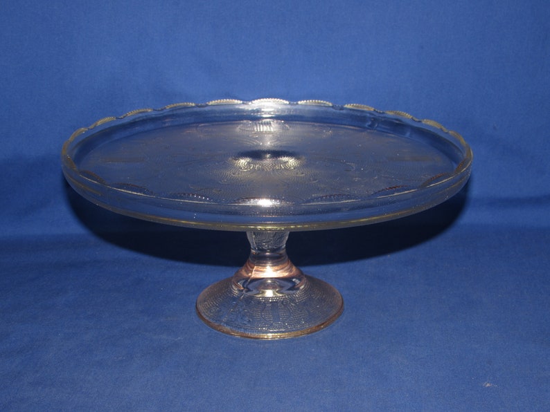 May include: A clear glass cake stand with a scalloped edge and a pedestal base. The stand has a delicate etched pattern.