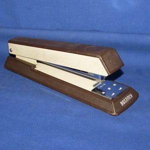 May include: A brown and beige stapler with the brand name "BOSTITCH" printed on the front. The stapler is made of metal and plastic and is set against a blue background. The stapler is open, ready for use.