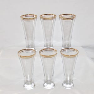 May include: Six clear glass pilsner glasses with a gold rim. The glasses are arranged in two rows of three, with a slightly flared shape. The gold rim adds a touch of elegance to the glassware.