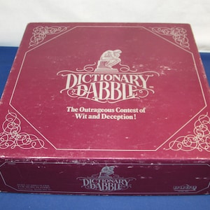 May include: A maroon-colored board game box with the title "Dictionary Dabble" in white script. The box features decorative corner designs and the tagline "The Outrageous Contest of Wit and Deception!"