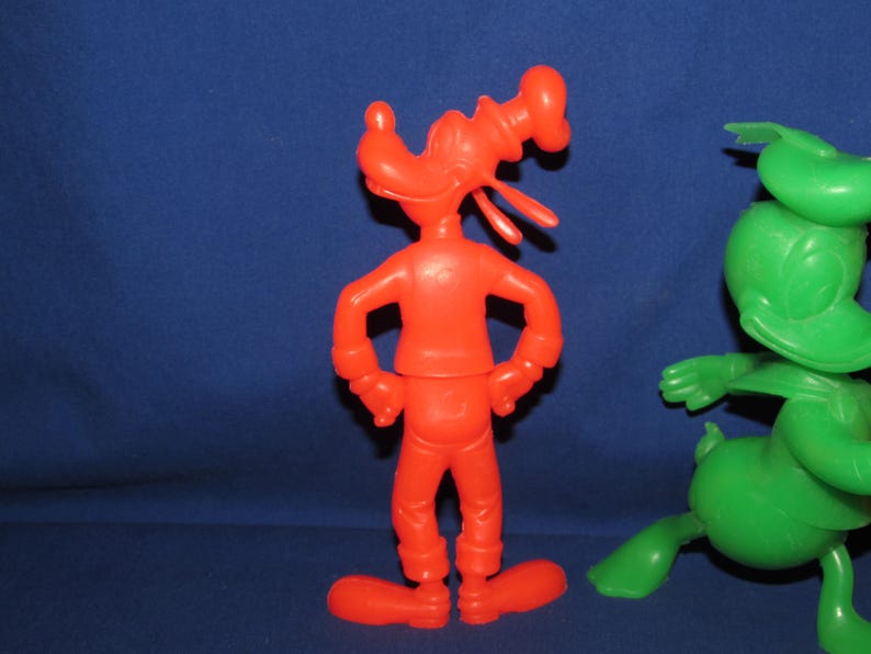 May include: Two plastic cartoon character figurines. One is red and is a caricature of a dog with a sailor hat. The other is green and is a caricature of a duck with a sailor hat.