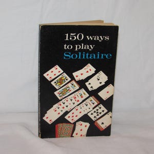 May include: A vintage book titled "150 ways to play Solitaire" with a black cover. The cover features the title in white and blue, along with illustrations of playing cards. The cards are arranged to appear as if they are falling.