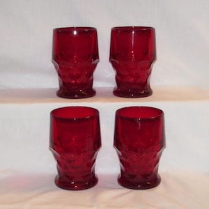 May include: Four ruby red drinking glasses. Each glass has a faceted design and a thick base. The glasses are arranged in two rows of two, against a white background. The glasses are empty.