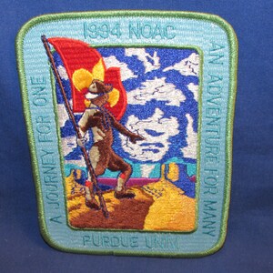 May include: A blue and green embroidered patch with a scout holding a flag. The text on the patch reads "1994 NOAC A Journey For One An Adventure For Many Purdue Univ."