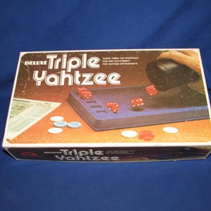 May include: A rectangular game box for Deluxe Triple Yahtzee featuring a blue dice tray, red dice, and white tokens. The box cover displays the game title, the text Three Times The Strategy, Fun And Excitement For Yahtzee Enthusiasts, and a score card.