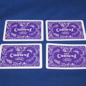 May include: Four playing cards with a purple and white design. Each card features the text "FIVE CROWNS" with a crown illustration. The cards are arranged on a blue surface.