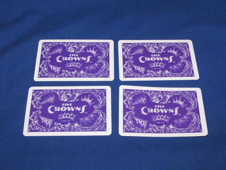 May include: Four playing cards with a purple and white design. Each card features the text "FIVE CROWNS" with a crown illustration. The cards are arranged on a blue surface.