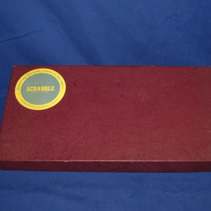 May include: A rectangular, maroon Scrabble game box with a circular label. The label is light green with gold lettering and a gold border. The text on the label reads "SCRABBLE" and "MANUFACTURED BY SELCHOW & RUGHTER CO. NEW YORK".