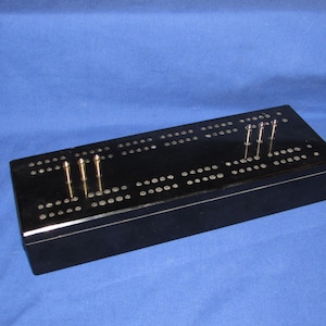 May include: A black cribbage board with metal pegs inserted. The board has rows of holes for scoring and is rectangular in shape. The pegs are silver and gold in color. The board is set against a blue background.