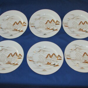 May include: Set of six white ceramic dinner plates with a gold rim and a gold design of a Japanese landscape with mountains, trees, and a bridge.