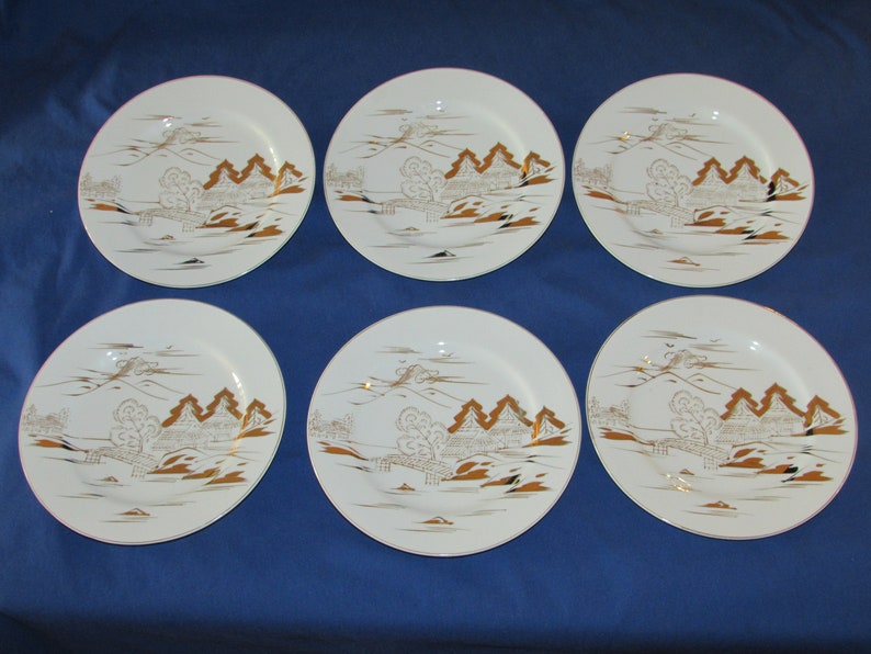 May include: Set of six white ceramic dinner plates with a gold rim and a gold design of a Japanese landscape with mountains, trees, and a bridge.