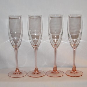May include: A set of four clear glass champagne flutes, each featuring a slender, tall bowl and a delicate pink-tinted stem and circular base. The glasses are arranged in a single row, showcasing their matching design and elegant, elongated shape.