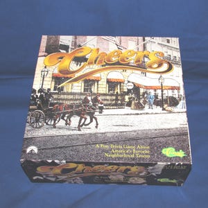 May include: A board game box titled "Cheers" with a vintage illustration of a tavern and horse-drawn carriage. The box features the game's title in gold lettering and the tagline "A Fun Trivia Game About America's Favorite Neighborhood Tavern."