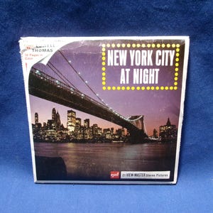 May include: A square View-Master packet featuring a night photograph of the Brooklyn Bridge and the New York City skyline. The text reads NEW YORK CITY AT NIGHT and 21 VIEW-MASTER Stereo Pictures. The packaging is white with a purple sky.
