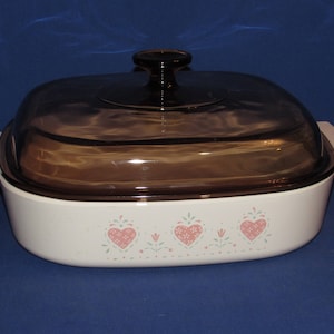 May include: White casserole dish with a brown glass lid. The dish has a decorative pattern of pink hearts and green floral accents. The lid has a black handle.