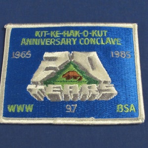 May include: A blue embroidered patch with white border, featuring the text "KIT-KE-HAK-O-KUT ANNIVERSARY CONCLAVE 1965 1985 WWW 97 BSA" and a silver 3D "20 YEARS" design with a green triangle and a brown beaver.