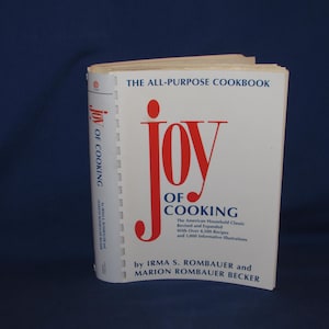 May include: A hardcover cookbook titled "Joy of Cooking" with a white cover and red lettering. The book is open, displaying the title and author's names. The spine reads "Joy of Cooking" vertically.