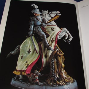 May include: A porcelain figurine of a knight on horseback slaying a dragon. The knight is wearing armor and a flowing cape, and the horse is white with gold accents. The dragon is green and has a chain around its neck.