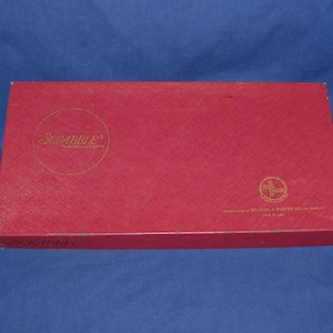 May include: A red Scrabble game box with gold lettering and a circular logo. The box is rectangular and has a textured surface. The game box is on a blue background.