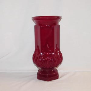 May include: A tall, ruby-red glass vase with a hexagonal body and a flared top. The vase features ornate, raised floral designs near the base. The base is a smaller, hexagonal shape. The vase is approximately 12 inches tall.