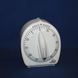 May include: A white Lux kitchen timer with a rounded, teardrop-shaped housing. The face features a circular dial with black numerals from 0 to 55, marked as MINUTE MINDER. A central rotating knob allows for setting time intervals.