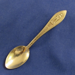 May include: A gold-colored teaspoon with a decorative design on the handle. The design features a person standing on a pedestal with a crown on their head. The handle is inscribed with the words "The Good Old Days".