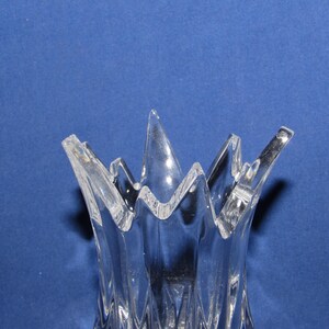 May include: Clear glass vase with a unique, jagged rim design. The vase has a textured, ridged body and a flared, starburst-like top. The transparent glass allows light to pass through, creating a play of shadows. The vase is set against a blue background.
