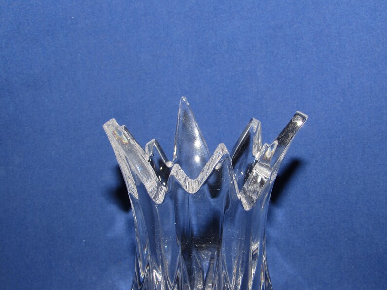 May include: Clear glass vase with a unique, jagged rim design. The vase has a textured, ridged body and a flared, starburst-like top. The transparent glass allows light to pass through, creating a play of shadows. The vase is set against a blue background.
