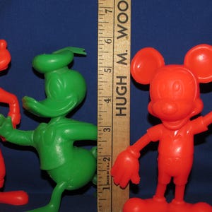 May include: Three vintage plastic cartoon character figurines. A red Goofy, a green Donald Duck, and a red Mickey Mouse. The figurines are standing on a wooden ruler with the text "HUGH M. WOOD" printed on it.