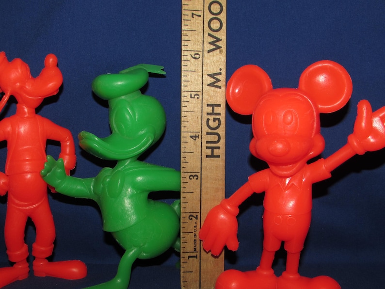 May include: Three vintage plastic cartoon character figurines. A red Goofy, a green Donald Duck, and a red Mickey Mouse. The figurines are standing on a wooden ruler with the text "HUGH M. WOOD" printed on it.