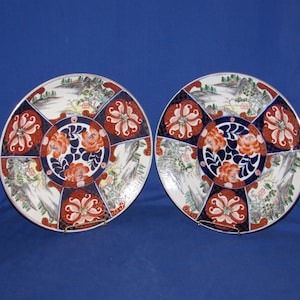 May include: Two decorative ceramic plates with intricate floral and landscape designs. The plates feature a central floral motif in orange and blue, surrounded by panels of landscapes and stylized flowers. The plates are on a blue background.