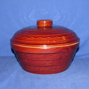 May include: A round, lidded, ceramic casserole dish in shades of brown and red. The dish features decorative horizontal ridges and floral patterns. The lid has a small, round handle. The dish is likely vintage and suitable for serving or storage.