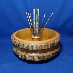 May include: A round wooden bowl with a natural bark edge featuring a central holder containing six silver-toned metal nut picks. The bowl has a smooth, light brown interior finish, while the exterior retains the rough, dark texture of tree bark.