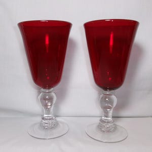 May include: A pair of matching wine glasses featuring deep red glass bowls and clear glass stems with a rounded decorative accent above a circular base. The glasses have a wide, flared rim and a smooth, polished finish throughout the structure.